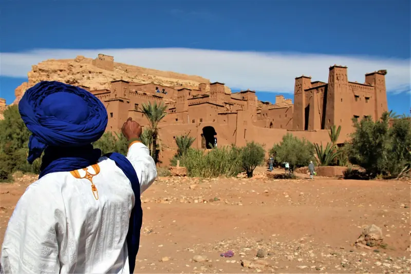 A guide in a blue turban pointing toward Ait Ben Haddou
